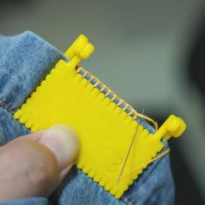 以下が含まれることがあります： 青いデニム生地に縫い目をつけるために使用される明るい黄色の縫製ゲージツール。ツールは、スカラップエッジと2つの小さなハンドルを備えた長方形の形状をしています。針と糸が見えます。