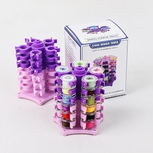Op de afbeelding: Een paarse en roze spoeltoren met meerdere niveaus, geschikt voor verschillende kleuren garen. De toren is ontworpen om tot 30 spoelen netjes te organiseren. Een doos met de tekst "STORE BOBBIN TOWER" is ook zichtbaar.