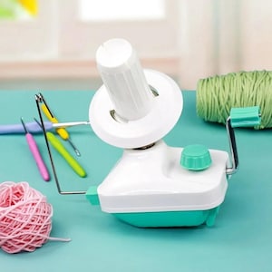 Op de afbeelding: Een witte en teal garenwinder met een handslinger, een witte spoel en een teal knop. Een roze bol garen en groen garen liggen naast de winder. Haaknaalden in roze, geel en groen zijn ook zichtbaar.