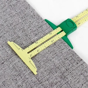 Puede incluir: Una herramienta de medición de costura deslizante de color amarillo y verde sobre una tela gris texturizada. La herramienta tiene un extremo en forma de T y un marcador deslizante con marcas métricas en centímetros, hasta 15 centímetros.