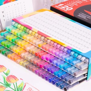 May include: A set of 120 colorful markers arranged in a box. The markers are in a rainbow of colors, from red to purple, with clear caps. The box has a label with the text "120 COLORS/PCS". A calendar and a box with the text "PAPER, ROCK, CERAMIC, GLASS, CANVAS" are in the background.