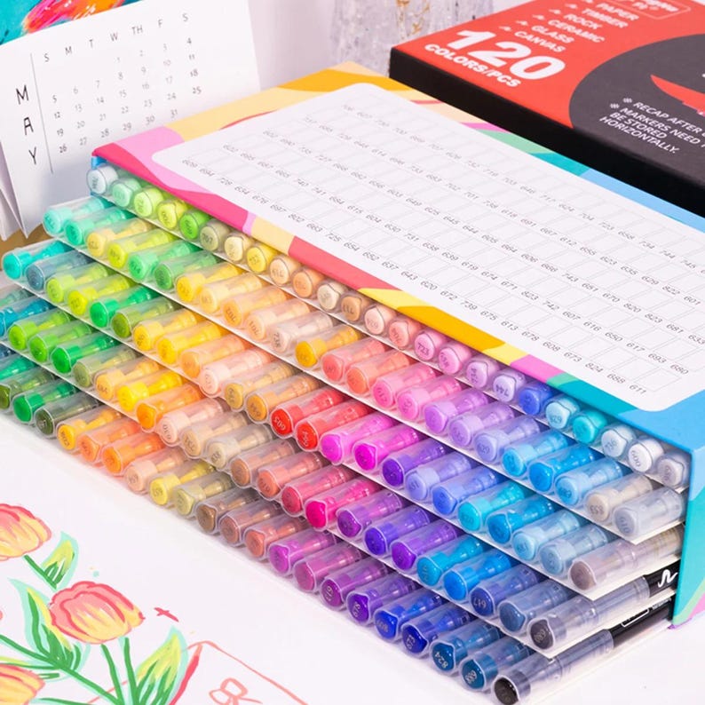 May include: A set of 120 colorful markers arranged in a box. The markers are in a rainbow of colors, from red to purple, with clear caps. The box has a label with the text "120 COLORS/PCS". A calendar and a box with the text "PAPER, ROCK, CERAMIC, GLASS, CANVAS" are in the background.