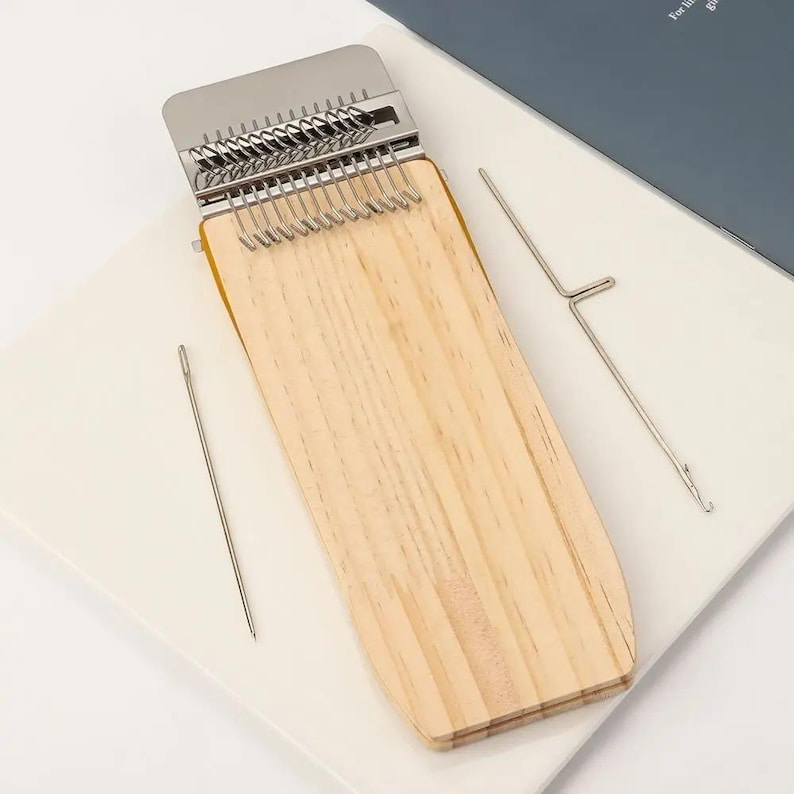 May include: A wooden kalimba with metal tines and a metal bridge. Two metal tools are included. The kalimba is light brown with a rectangular shape and rounded edges. The tines are silver. The tools are silver and have a needle-like appearance.