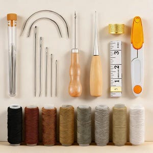 Peut inclure: Ensemble d'outils et de fournitures pour le travail du cuir. On y trouve des aiguilles, des alènes, des bobines de fil de différentes couleurs, un dé à coudre, un mètre ruban et un petit couteau pliant orange et blanc. Le tout est disposé sur une surface beige clair.