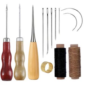 May include: A leathercrafting kit featuring various needles, awls with wooden handles, spools of black and brown thread, and a gold thimble. The tools are arranged on a white background, showcasing the essential components for leatherwork.