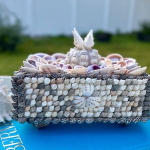 May include: A decorative box made entirely of seashells. The box is square and has a lid with a seashell sculpture of a bird. The box is sitting on a blue book.