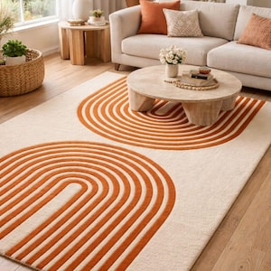 May include: A cream-colored area rug with two large, orange arched designs. The rug is placed in a living room setting with a beige sofa, wooden coffee table, and a window with natural light.