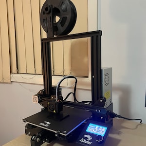 May include: A black 3D printer with a filament spool holder, a blue LCD screen, and the word "Ender" printed on the front.