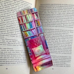 Puede incluir: Un marcapáginas de acuarela con un sillón rosa con un cojín, una estantería llena de libros coloridos y una cita de un libro. La cita dice: "Sigue siendo una habitación, que puedes mirar."