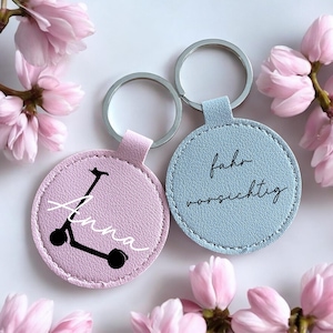 Könnte beinhalten: Zwei runde Schlüsselanhänger, einer rosa und einer blau, mit silbernen Schlüsselringen. Der rosa Schlüsselanhänger zeigt eine schwarze Rollergrafik und den Namen "Anna" in weißer Schrift. Der blaue Schlüsselanhänger hat den Schriftzug "fahr vorsichtig" in schwarzer Schrift.