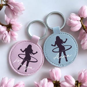 Könnte beinhalten: Zwei personalisierte Schlüsselanhänger mit einem Hula-Hoop-Silhouette-Design. Ein Schlüsselanhänger ist hellrosa, der andere hellblau. Jeder hat einen silbernen Schlüsselring und einen Namen in weißer Schrift. Rosa Blüten umgeben die Schlüsselanhänger.