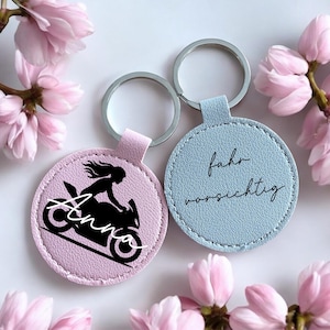 Op de afbeelding: Twee ronde sleutelhangers met zilveren ringen. Eén is lichtroze met een zwarte motorfiets- en rijderafbeelding, de naam "Anna" en een lichtroze bandje. De andere is lichtblauw met de woorden "fahr vorsichtig" en een lichtblauw bandje. Roze bloemen omringen de sleutelhangers.