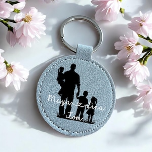 Può includere: Un portachiavi rotondo azzurro con un anello argentato. Il portachiavi presenta una silhouette nera di una persona che tiene in braccio due bambini, con altri due bambini nelle vicinanze. I nomi "Maya", "Dina" e "Tom" sono scritti in corsivo bianco.