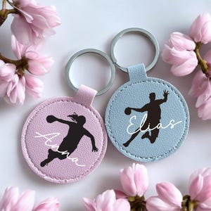 Könnte beinhalten: Zwei personalisierte Schlüsselanhänger mit Sportmotiv. Einer ist hellblau, der andere lavendelfarben. Jeder Schlüsselanhänger zeigt eine Silhouette einer Person beim Sport und einen Namen in Schreibschrift. Rosa Blüten umgeben die Schlüsselanhänger.