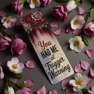 Könnte beinhalten: Ein rechteckiger Lesezeichen mit dem Text "You HAD ME at Trigger Warning" und einem floralen Design. Das Lesezeichen ist von rosa Rosen, weißen Blumen und Blütenblättern auf einer grauen Oberfläche umgeben.