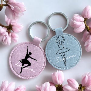 Könnte beinhalten: Zwei runde Schlüsselanhänger mit Balletttänzerinnen-Silhouetten. Einer ist hellrosa mit einer schwarzen Tänzerin und dem Namen "Liana" in weißer Schrift. Der andere ist hellblau mit einer schwarzen Tänzerin und dem Namen "Samira". Silberne Schlüsselringe.