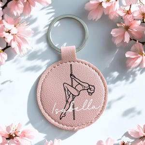 Può includere: Un portachiavi rosa rotondo con un anello argentato. Il portachiavi presenta un disegno a tratto nero di una persona che balla alla sbarra, con il nome "Isabella" in corsivo bianco. Fiori rosa sullo sfondo.