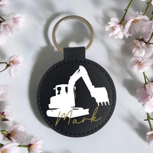 Könnte beinhalten: Runder Schlüsselanhänger aus schwarzem Leder mit silbernem Schlüsselring. Er zeigt ein weißes Bagger-Design und den Namen "Mark" in goldener Schrift. Der Schlüsselanhänger ist von rosa Blüten umgeben.