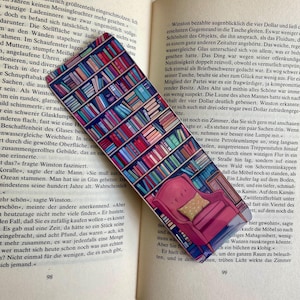 Könnte beinhalten: Ein roter Sessel mit einem Kissen steht vor einem Bücherregal, das mit bunten Büchern gefüllt ist. Das Lesezeichen ist ein rechteckiges Glas mit einem Bücherregal-Design.
