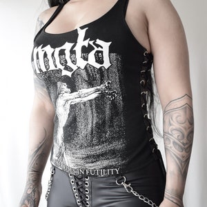 May include: Black tank top with a white graphic print of a band name and a graphic image. The tank top has lace-up detailing on the sides. The model is wearing black pants with a lace-up detail and a chain.