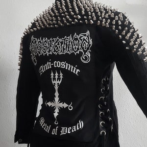 Op de afbeelding: Zwart denim jack met zilveren studs op de schouders. Het jack heeft een wit geborduurd ontwerp op de rug dat "Desecration", "Anti-cosmic" en "Metal of Death" zegt.