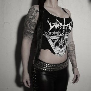 Puede incluir: Un top negro sin mangas con un gráfico blanco de un logotipo de banda que dice "Watain" y "Endless Darkness". El top está recortado y tiene un aspecto desgastado. La persona que lleva el top tiene el pelo negro y tatuajes en los brazos.