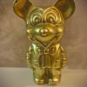 May include: A gold-colored figurine of a cartoon mouse wearing a suit and bow tie. The mouse has large, round ears and a smiling expression. The figurine is standing upright on a white surface.