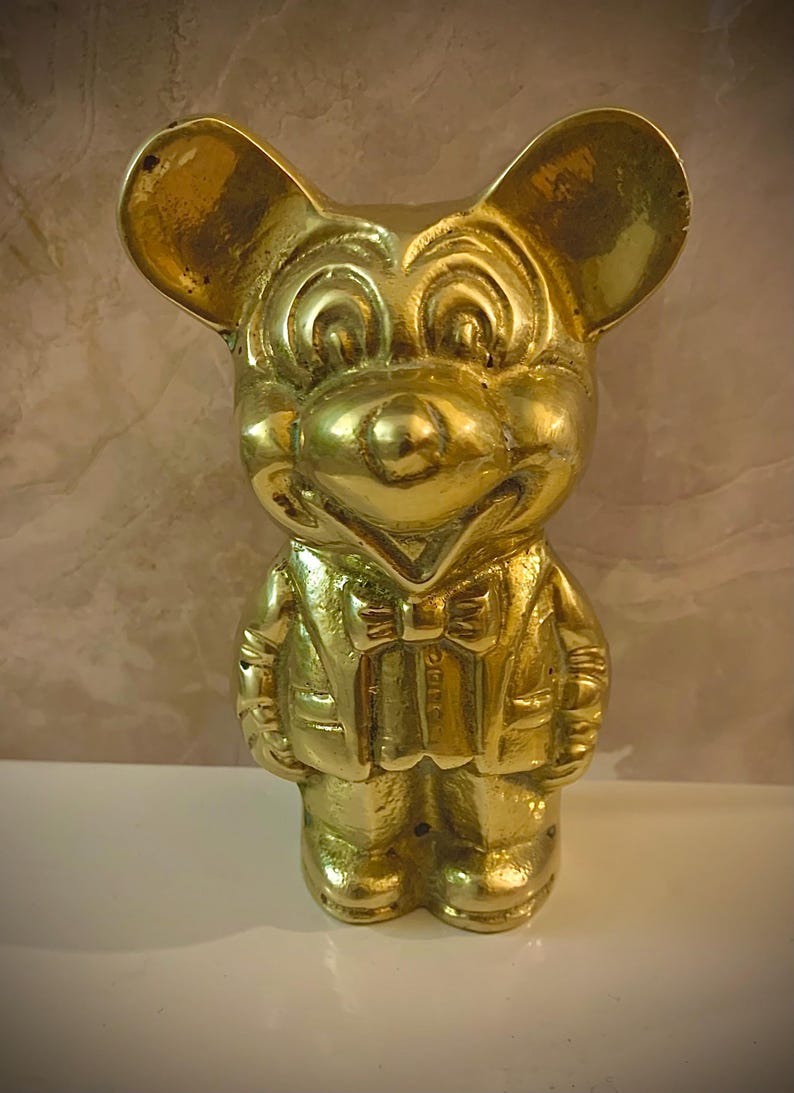 May include: A gold-colored figurine of a cartoon mouse wearing a suit and bow tie. The mouse has large, round ears and a smiling expression. The figurine is standing upright on a white surface.