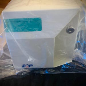Peut inclure: Une boîte de rangement rectangulaire blanche enveloppée dans un emballage plastique transparent. L'avant présente une zone d'étiquette rectangulaire turquoise, une serrure à clé argentée circulaire à droite et le texte EXP en bleu et rouge.
