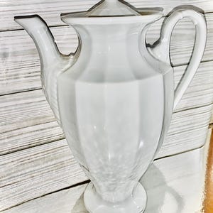 May include: A white ceramic coffee pot with a lid and handle. The pot has a textured design on the side and a decorative finial on the lid. The pot is on a white surface.