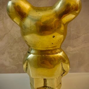 May include: A solid brass figurine in the shape of a bear. The bear is standing upright with its arms at its sides. The bear has a smooth, polished surface and a golden color. The figurine is a decorative item.