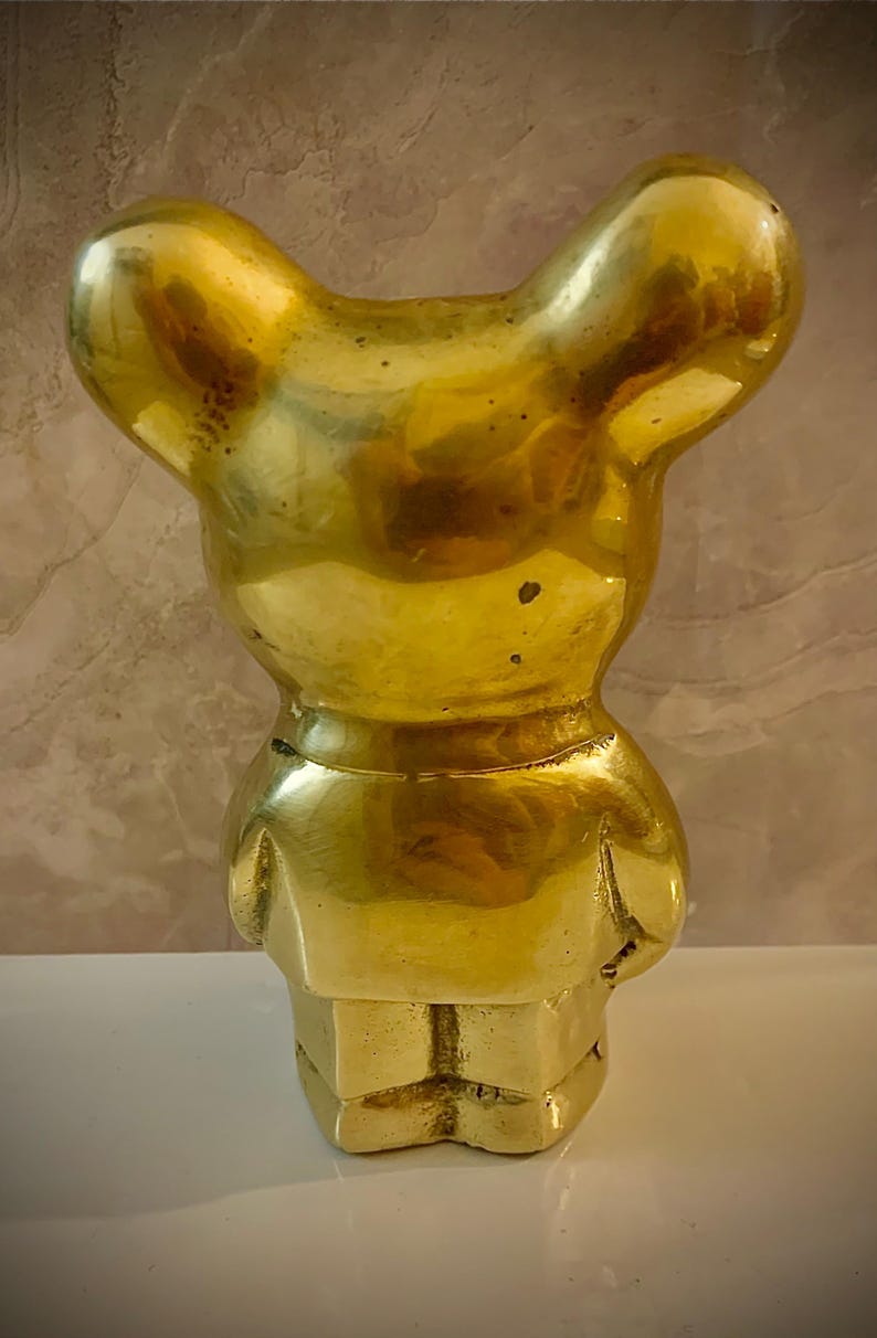 May include: A solid brass figurine in the shape of a bear. The bear is standing upright with its arms at its sides. The bear has a smooth, polished surface and a golden color. The figurine is a decorative item.