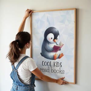 May include: A framed print featuring a watercolor illustration of a penguin reading a book. The penguin is gray, black, and white with orange feet and a pink book. The text "COOL KIDS read books" is below the penguin.