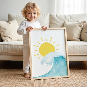 May include: A framed art print depicting a yellow sun and a blue watercolour wave. The artwork is held by a young child. The frame is made of light-coloured wood. The background includes a beige sofa and a neutral-coloured rug.