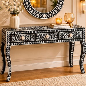 May include: A black console table featuring intricate white floral and vine inlay patterns across its rectangular top, three front drawers, and four curved legs. Each drawer is equipped with a round, white, flower-shaped knob for opening.