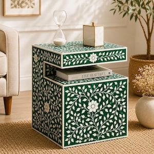 Puede incluir: Una mesa auxiliar con estampado floral verde y blanco con un diseño de dos niveles. La mesa presenta una forma cuadrada con intrincadas incrustaciones de hojas y flores. Un libro con el texto "LIVE BEAUTIFUL" está en el estante inferior. Un reloj de arena y una caja decorativa están encima.