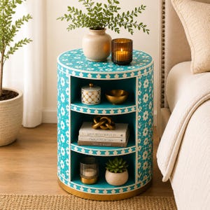 May include: A round, turquoise side table with white floral patterns and gold trim. The table has three shelves displaying decorative items like books, a gold bowl, a candle, and a small succulent plant. A vase with greenery and a candle holder sit on top.