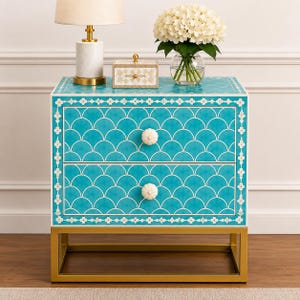 May include: A two-drawer nightstand featuring a vibrant turquoise finish with a white scalloped pattern and floral border inlay. The piece includes two round, fluted white knobs and is supported by a rectangular gold-toned metal frame base.