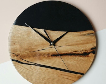 Puede incluir: Un reloj redondo de madera con detalles de resina epoxi negra. La esfera del reloj está hecha de madera natural con un patrón de veta único. Las manecillas son negras y el reloj tiene un diseño minimalista.