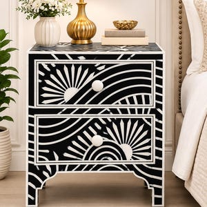 May include: A two-drawer bedside table featuring a bold black and white geometric pattern of radiating sunbursts and concentric arcs. The rectangular piece has a flat top surface, four short legs with arched cutouts, and two round white knobs.