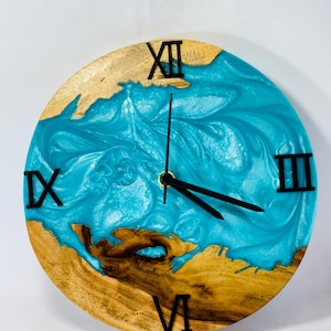 May include: A round wooden clock with a blue resin face. The clock has Roman numeral markings and black hands. The wood is a light brown colour with darker brown streaks.