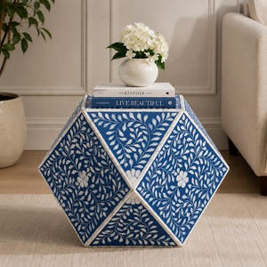 May include: A blue and white hexagonal side table with a floral leaf design. The table has a vase of white flowers, and two books on top. The books are titled "THE KINFOLK HOME" and "LIVE BEAUTIFUL".