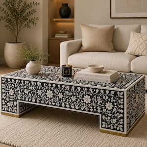 May include: A black and white floral patterned coffee table with gold accents. The rectangular table is in front of a white sofa with beige pillows. A candle, books, and a small bowl are on the table. The room has a neutral color palette.