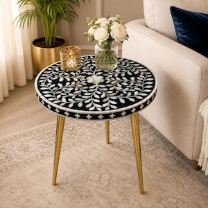 Pode incluir: Uma mesa lateral redonda com um tampo preto incrustado com um padrão floral branco. A mesa tem três pernas douradas. Um vaso de rosas brancas e um castiçal dourado estão sobre a mesa.