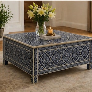 May include: Square coffee table with a navy blue and white floral pattern. The table has a decorative design on the top and sides. A vase of white flowers, a candle, and a book are on the table.