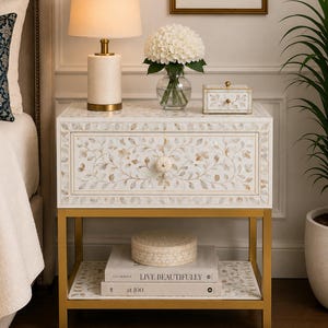 May include: A white bedside table featuring an intricate floral mother-of-pearl inlay, a single drawer with a round knob, and a gold metal frame. The lower shelf holds two stacked books titled "LIVE BEAUTIFULLY" and "at 100" with a round box on top.