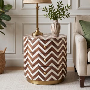 Può includere: Un comodino cilindrico a tre cassetti con un motivo a chevron marrone e bianco sull'esterno. Il mobile include tre piccoli pomelli rotondi color oro e una base circolare color oro. La superficie è liscia e rifinita.