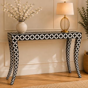 May include: A rectangular console table featuring a black and white geometric pattern of interlocking circles. The table has four curved legs and a flat top surface. A white ribbed vase with white flowers and a gold lamp sit on the table.