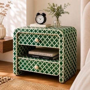 Puede incluir: Una mesita de noche de color verde oscuro con un patrón de cuatrifolio blanco, que cuenta con dos cajones con pomos blancos redondos. La superficie superior sostiene un despertador analógico negro y un jarrón de cristal con follaje verde.