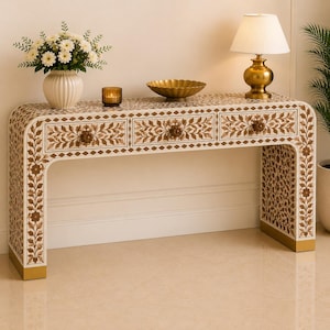 May include: A rectangular console table featuring an intricate floral inlay pattern in brown and cream. The table has three drawers with round, dark brown floral-shaped knobs and a curved frame design with gold-toned metal base accents.